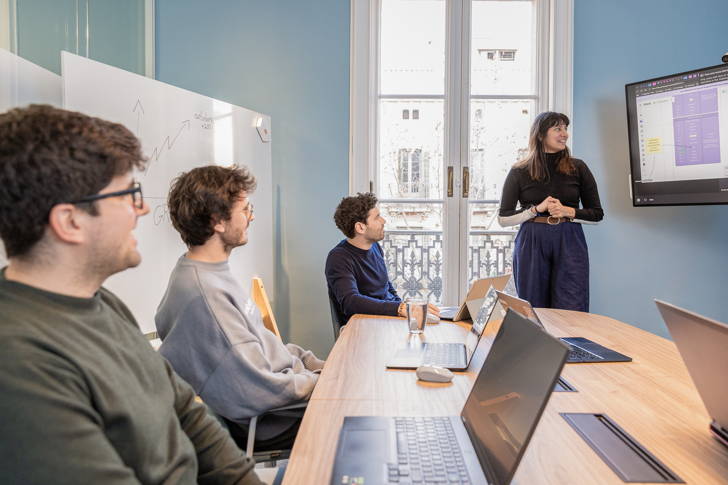 People at a working meeting, sitting on a big desk. One woman is standing presenting in front of a screen.