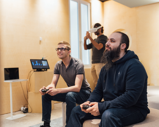 Two men playing a video game and another man in the background with VR glasses