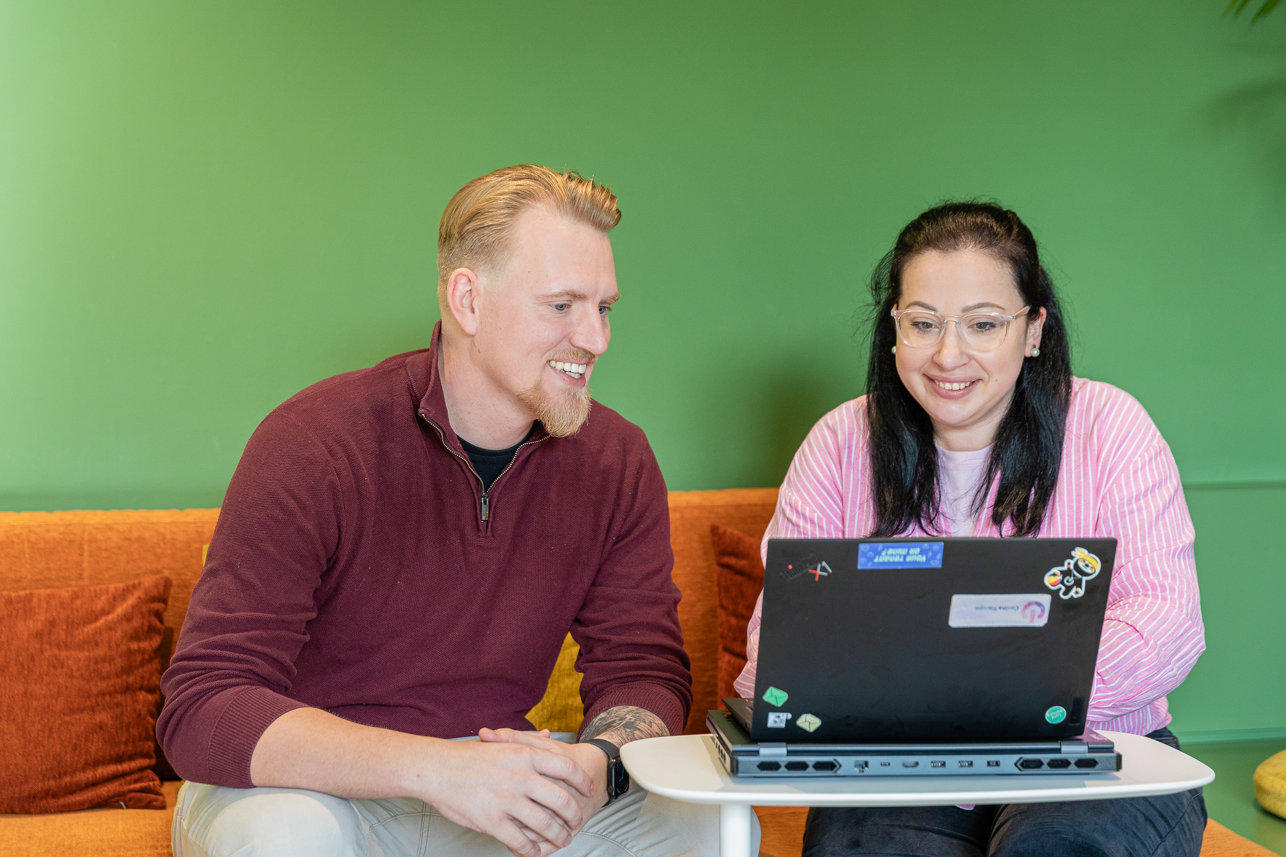 A men and a woman sitting in front of a laptop smiling. Green wall and orange sofa.