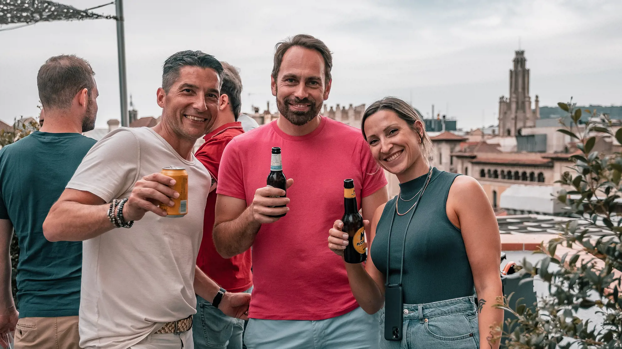Two men and a woman on a terrace toasting to the camera with a beer